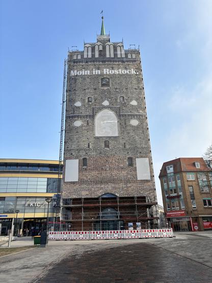 Am Stadttor Kröpeliner Tor in Rostock steht ein Gerüst in voller Höhe, das mit einer Plane mit einem Foto der Fassade des Tors verkleidet ist.